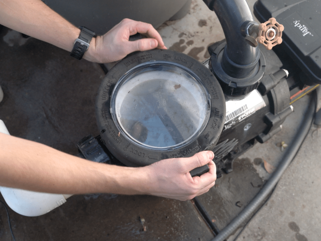 A person's hands unscrew the circular, transparent lid on a pool pump filter housing.