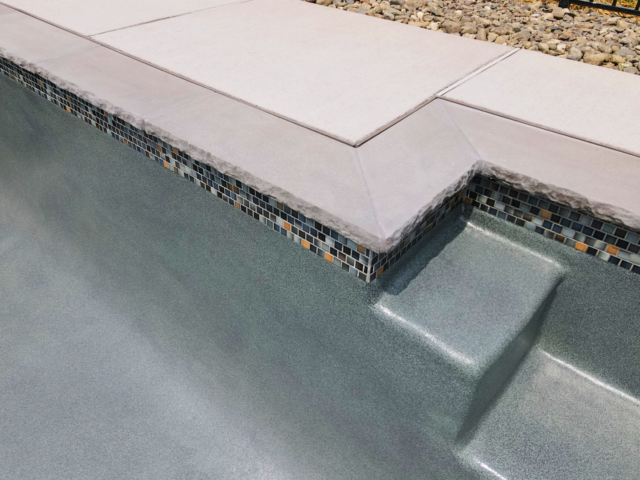 A close-up view of the interior and coping of a swimming pool. The pool's interior surface is a dark gray color with a speckled texture. A band of small, iridescent mosaic tiles in shades of blue, gray, and orange lines the top of the pool's wall. The coping is a light gray stone with a rough, chiseled edge, and the surrounding deck is made of smooth, light-colored concrete pavers.