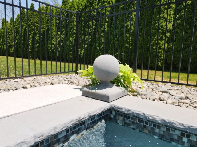 A close-up, low-angle shot of the corner of a swimming pool. The pool's coping is made of gray stone with a rough, chiseled edge, and the interior is lined with small, iridescent blue, gray, and brown mosaic tiles. On the patio, there is a gray concrete piece with a spherical top.