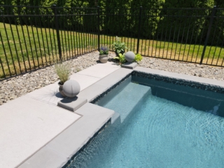 A wide-angle, eye-level shot of the corner of a swimming pool with built-in steps. The pool is lined with small mosaic tiles, and the coping is made of gray stone with a textured, chiseled edge. The patio is a light gray concrete, and a black metal fence encloses the area.