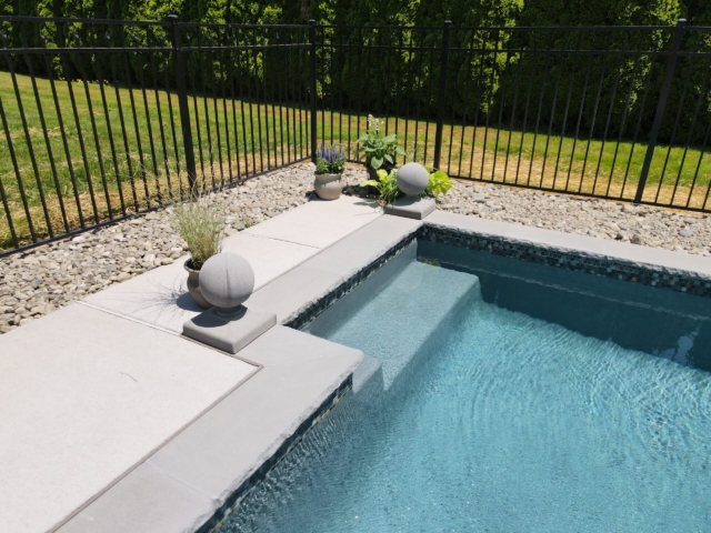 A wide-angle, eye-level shot of the corner of a swimming pool with built-in steps. The pool is lined with small mosaic tiles, and the coping is made of gray stone with a textured, chiseled edge. The patio is a light gray concrete, and a black metal fence encloses the area.