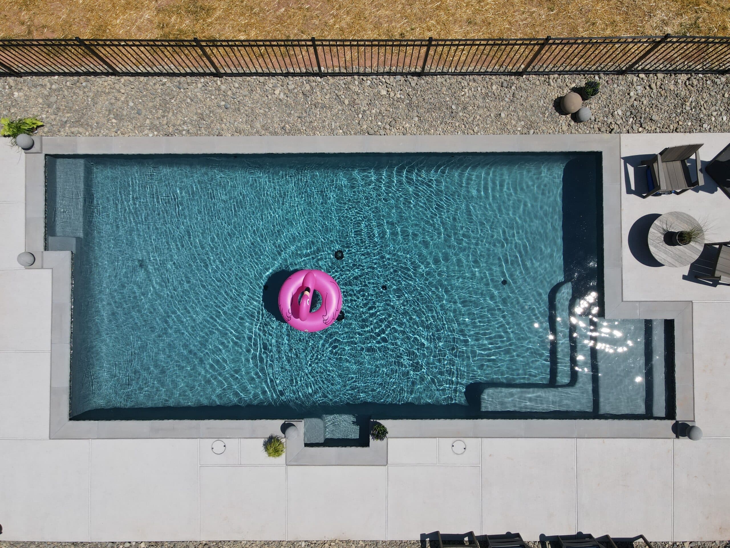 An overhead aerial view of a rectangular, dark-bottomed swimming pool with a hot pink flamingo inner tube floating in the center. The pool has a light gray concrete deck with built-in steps at one end. A dark gray coping surrounds the pool. There are some lounge chairs and small tables on the deck. A border of small, gray river rocks and a black metal fence surrounds the entire area.