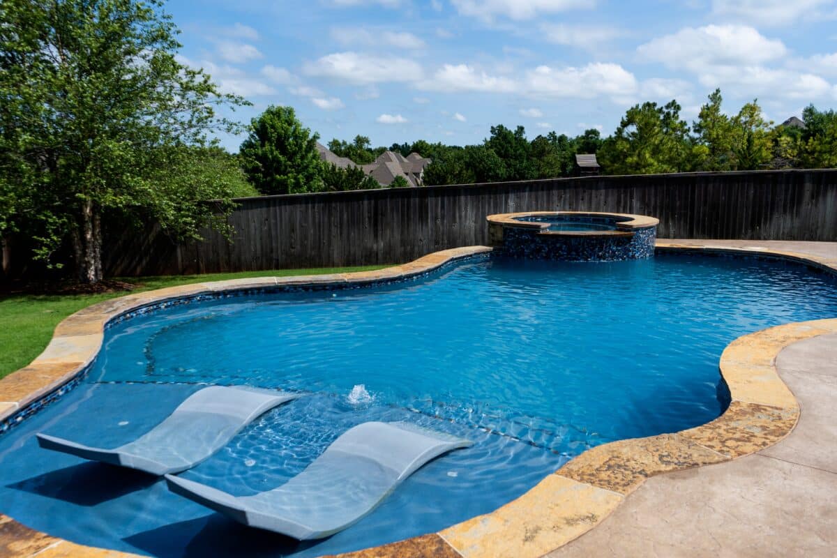 A sunny backyard with a large, freeform swimming pool and an integrated hot tub. The pool has a deep blue color, a built-in sun shelf with two submerged lounge chairs, and a small water fountain feature. The hot tub is circular and lined with dark blue mosaic tiles. The pool's coping is made of large, textured, sandy-colored stone slabs.