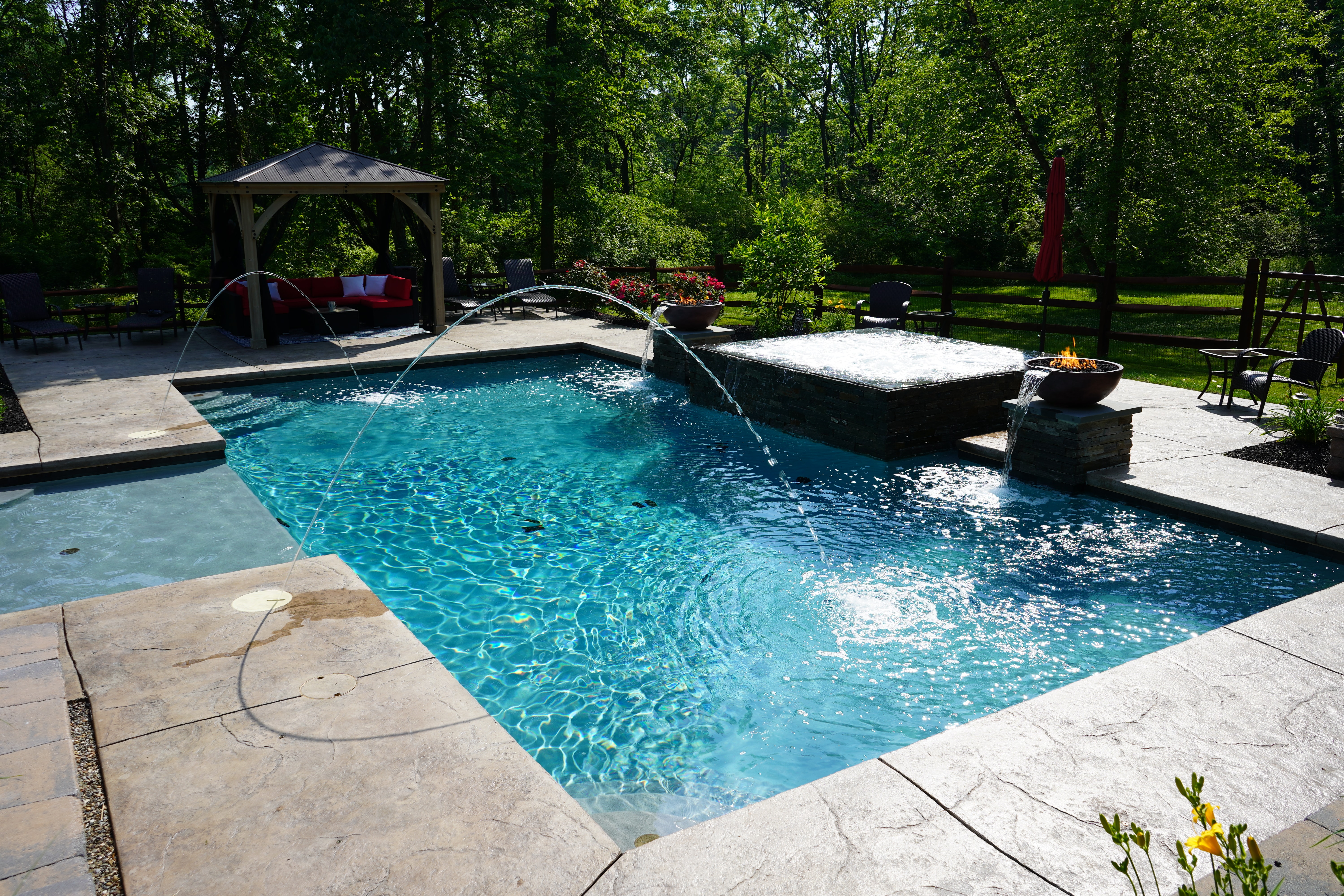 A beautifully landscaped backyard with a rectangular swimming pool and an integrated hot tub. The hot tub is clad in decorative stone. On either side of the hot tub are two stone columns, each topped with a large, bowl-like feature from which water spills into the pool. The bowls also have a fire feature with flames burning. In the background, there is a brown wooden fence, and a gazebo with red cushions is visible at the left. The yard is surrounded by lush green trees.
