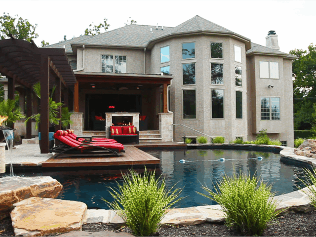 A luxurious backyard with a large swimming pool. The pool is kidney-shaped with a rope and buoys across the middle. A small, square deck extends into the pool. Two fire features are on opposite sides of the pool: one is a large bowl on a pedestal, and the other is a small fire pit surrounded by rocks.