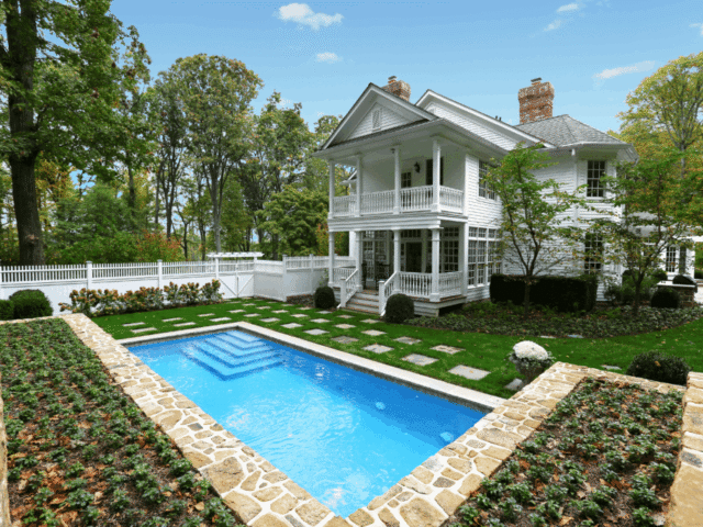 A beautiful two-story white house with a columned porch and a second-story balcony. In the foreground, a swimming pool is surrounded by a well-manicured lawn with stepping stones leading to the house. The pool is bordered by a stone retaining wall with low-growing plants.