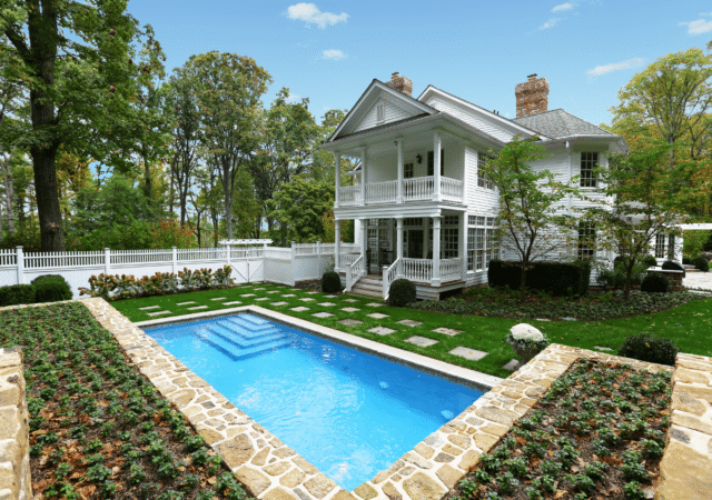 A beautiful two-story white house with a columned porch and a second-story balcony. In the foreground, a swimming pool is surrounded by a well-manicured lawn with stepping stones leading to the house. The pool is bordered by a stone retaining wall with low-growing plants.