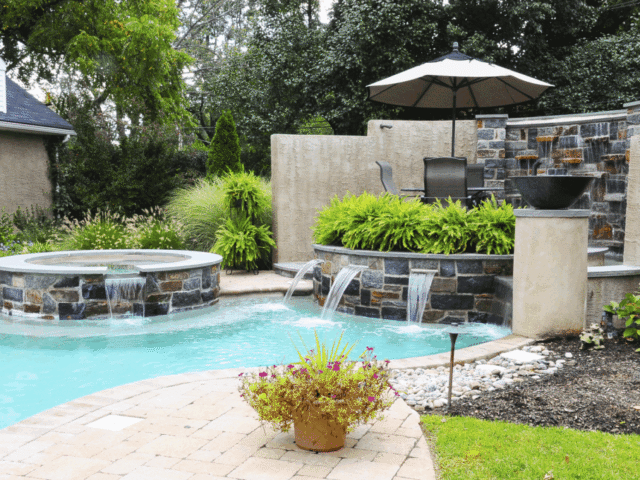 A ground-level shot of a backyard pool area with a detailed water feature. The circular hot tub on the left has a stacked stone exterior and a small waterfall flowing into the light-blue pool. On the right, a large, multi-level water feature is built with stacked stone and a textured wall. Water cascades from a wall of stone into a lower basin with three spouts.
