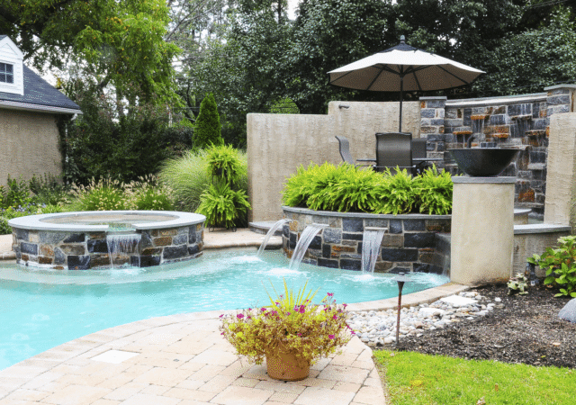 A ground-level shot of a backyard pool area with a detailed water feature. The circular hot tub on the left has a stacked stone exterior and a small waterfall flowing into the light-blue pool. On the right, a large, multi-level water feature is built with stacked stone and a textured wall. Water cascades from a wall of stone into a lower basin with three spouts.
