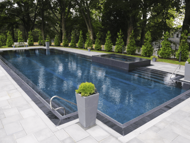 A high-angle view of a large, modern rectangular swimming pool. The pool has a dark interior. A raised hot tub with dark tile siding is visible at the far end, also overflowing into the pool. The pool deck is made of light-colored pavers, and the pool's edge features a dark, continuous grate system.