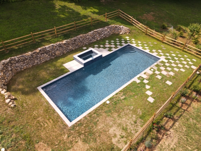 An aerial view of a rectangular, dark-bottom swimming pool with a connected square hot tub. The pool is surrounded by a light grey coping. The surrounding lawn is a mix of green grass and a checkerboard pattern of light-colored pavers.