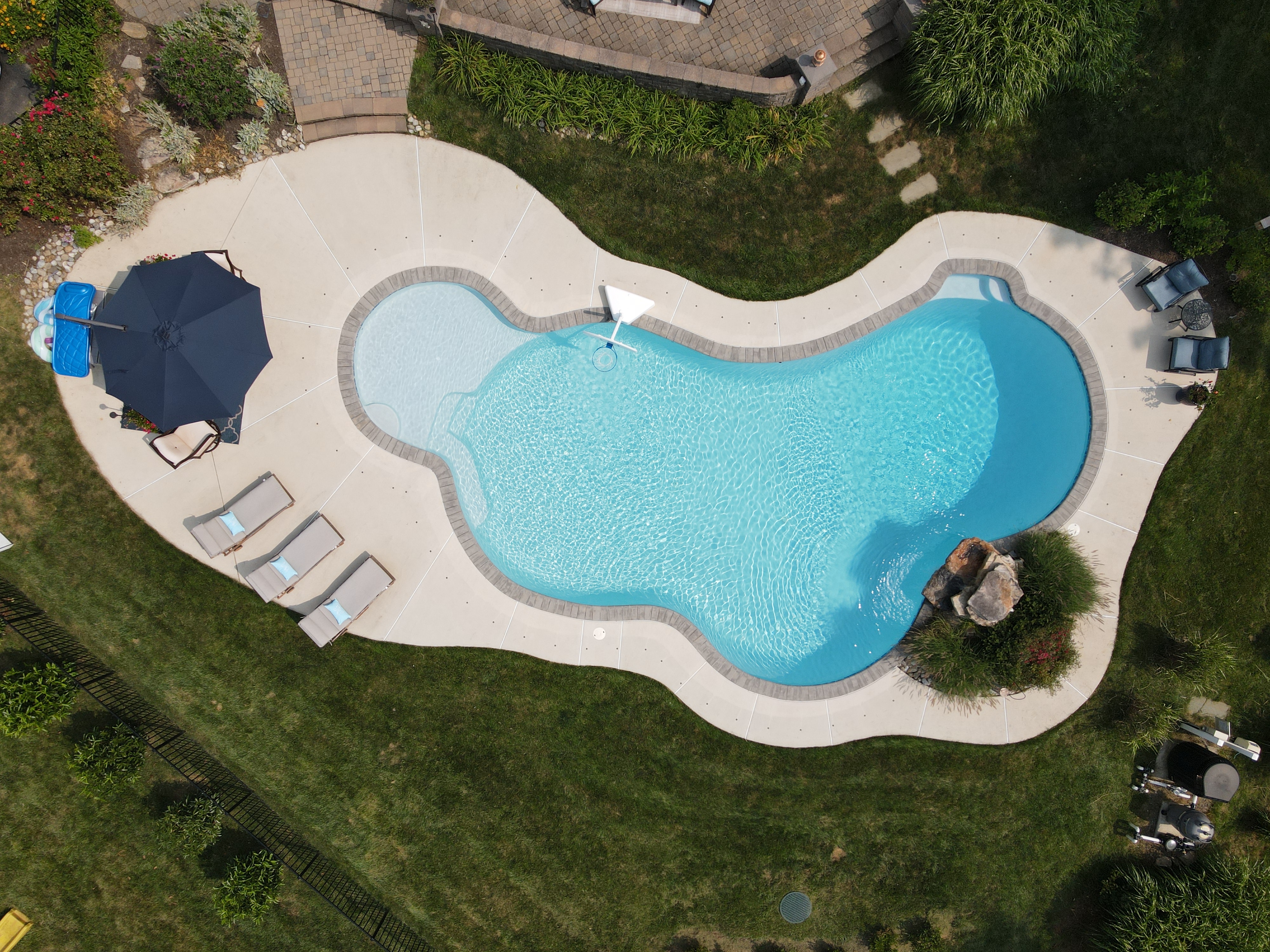 An aerial view of a bean-shaped swimming pool with a light blue bottom. The pool has a large sun shelf and is surrounded by a light-colored concrete deck. A large, dark blue umbrella provides shade for a few chairs. A rock feature with a waterfall is at the opposite end of the pool.