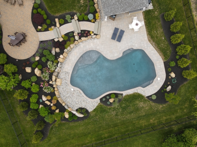 An aerial view of a backyard with a freeform swimming pool and a small, attached pergola. The pool has a dark blue interior and is surrounded by a light-colored paver deck. A set of stepping-stone stairs leads up a small, landscaped hill.