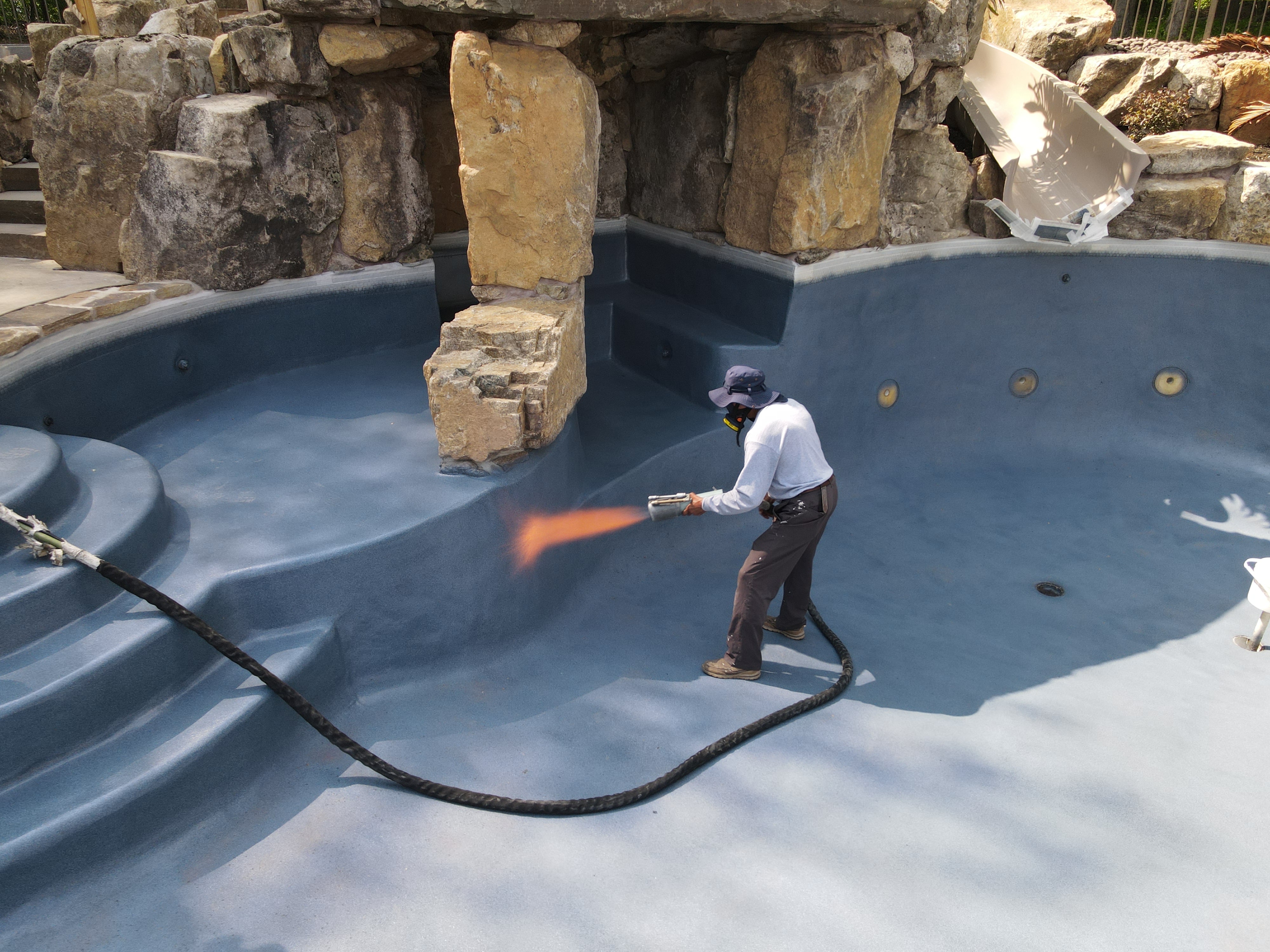 A worker wearing a sun hat and a white shirt sprays a coating onto the surface of an empty swimming pool. The pool has several steps and a large rock formation.