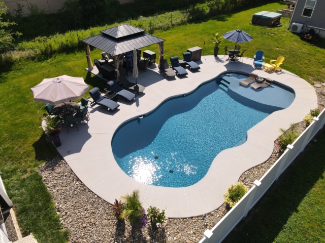 An aerial view of a backyard swimming pool with a unique, curvy shape and a sun shelf at one end. The pool has a dark blue interior and a light-colored concrete deck. On the sun shelf, there are two lounge chairs.