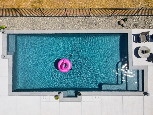 An aerial view of a rectangular swimming pool with a bright pink flamingo inner tube floating in the deep blue water. The pool has a sun shelf and steps at both ends and is surrounded by a light-colored concrete patio with a few pieces of patio furniture.