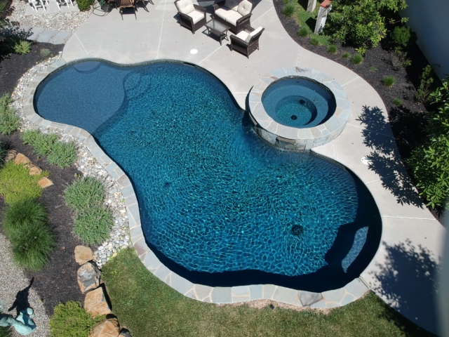 An overhead view of a custom-shaped backyard swimming pool and spa. The pool has a dark interior and is outlined by a natural stone coping. The spa, which is circular, is connected to the main pool and also has stone coping. The pool deck is a light-colored, curved concrete slab.