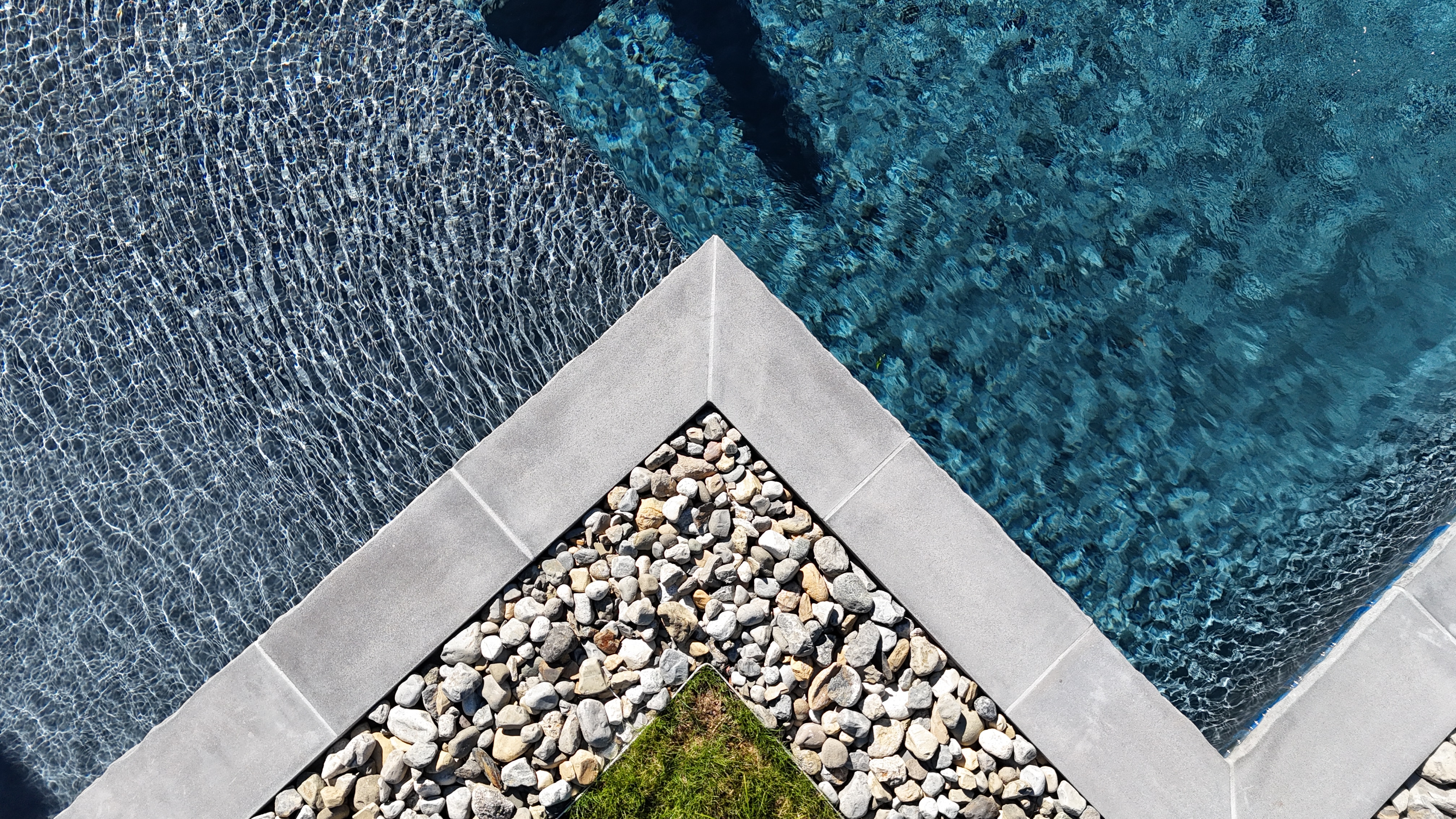 An overhead shot of the corner of a swimming pool. The pool's coping is a dark gray stone, creating a sharp angle. The pool has deep blue water. The deck area is a mix of small, light-colored river stones and a patch of green grass, creating a modern and natural look.