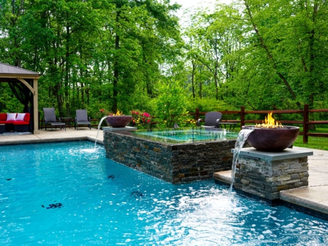 A swimming pool and hot tub are set in a lush green backyard, surrounded by trees. The hot tub is clad in stone and has two large bowls on either end that serve as fire and water features.