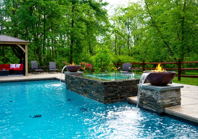 A swimming pool and hot tub are set in a lush green backyard, surrounded by trees. The hot tub is clad in stone and has two large bowls on either end that serve as fire and water features.