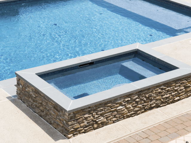 A rectangular, raised hot tub with a stone base and gray coping is next to a swimming pool. The water in the hot tub is a light blue, and steps are visible leading into it. The hot tub and pool are surrounded by a beige concrete patio.