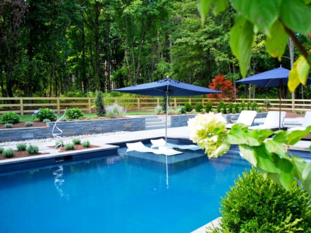 A poolside view of a modern swimming pool and patio area. The pool has a dark interior and features a shallow sun shelf with an in-pool lounge chair and a large blue patio umbrella. The pool area is surrounded by a light-colored patio and a retaining wall made of stacked stone, which supports a landscaped garden bed.