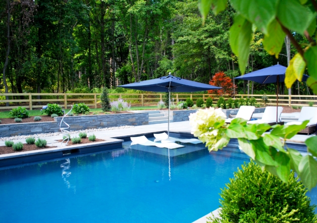 A poolside view of a modern swimming pool and patio area. The pool has a dark interior and features a shallow sun shelf with an in-pool lounge chair and a large blue patio umbrella. The pool area is surrounded by a light-colored patio and a retaining wall made of stacked stone, which supports a landscaped garden bed.