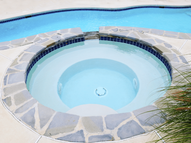 A round hot tub with light blue water is set into a concrete patio. The tub is surrounded by gray flagstones, with a small waterfall feature connecting it to a rectangular swimming pool behind it. The swimming pool has a dark blue tile border.