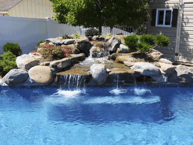 A waterfall feature with large rocks and lush green plants cascades into a swimming pool. In the background, a light yellow shed with a window and a white fence are visible.