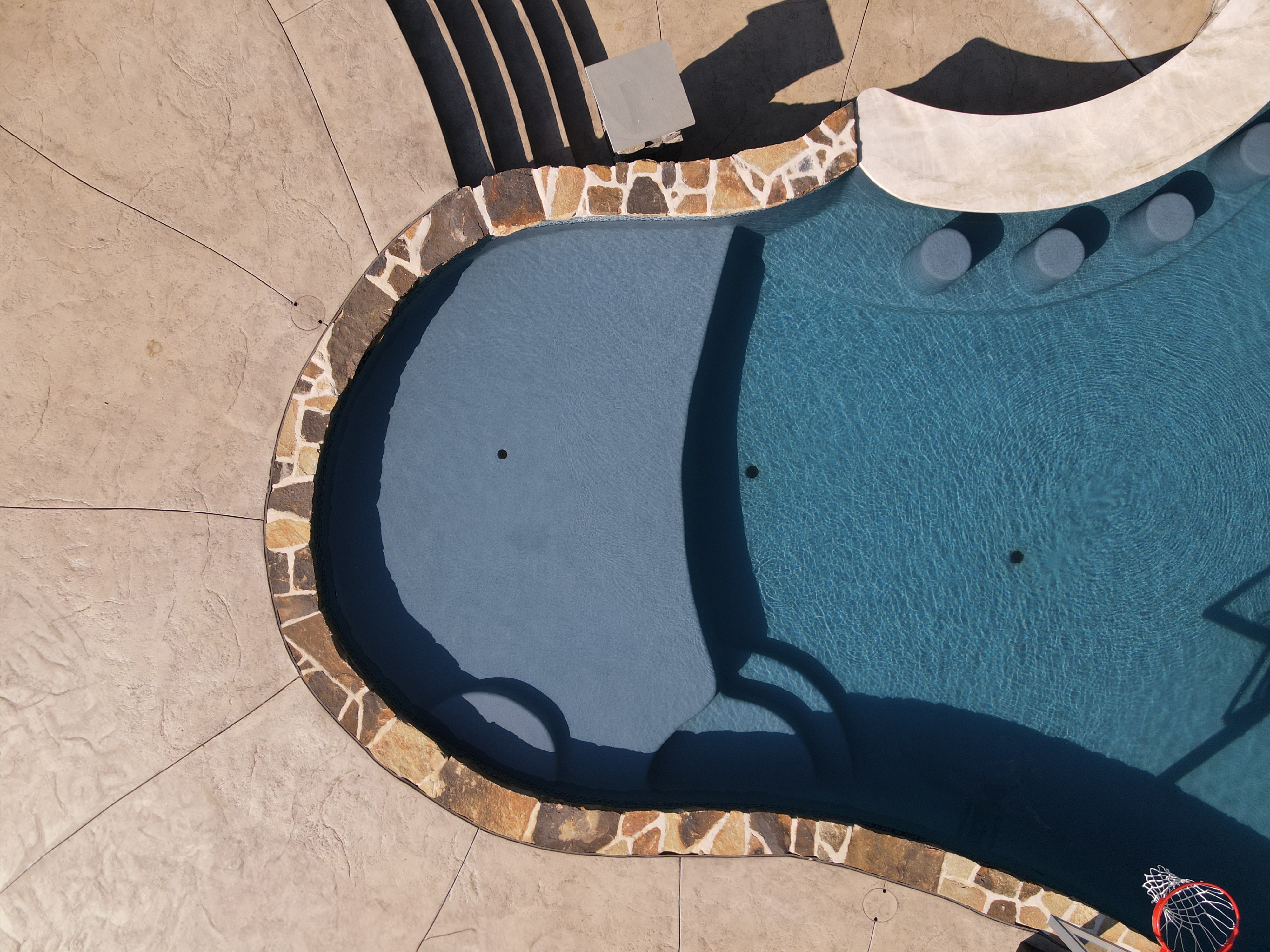 An overhead shot of a swimming pool sun shelf. The pool has a dark blue liner, and its edges are lined with a decorative, multi-toned stone coping. The surrounding deck is a light brown, stamped concrete. In the shallow end of the pool, there are built-in stools, and a small, raised platform with a lighter-colored stone border.