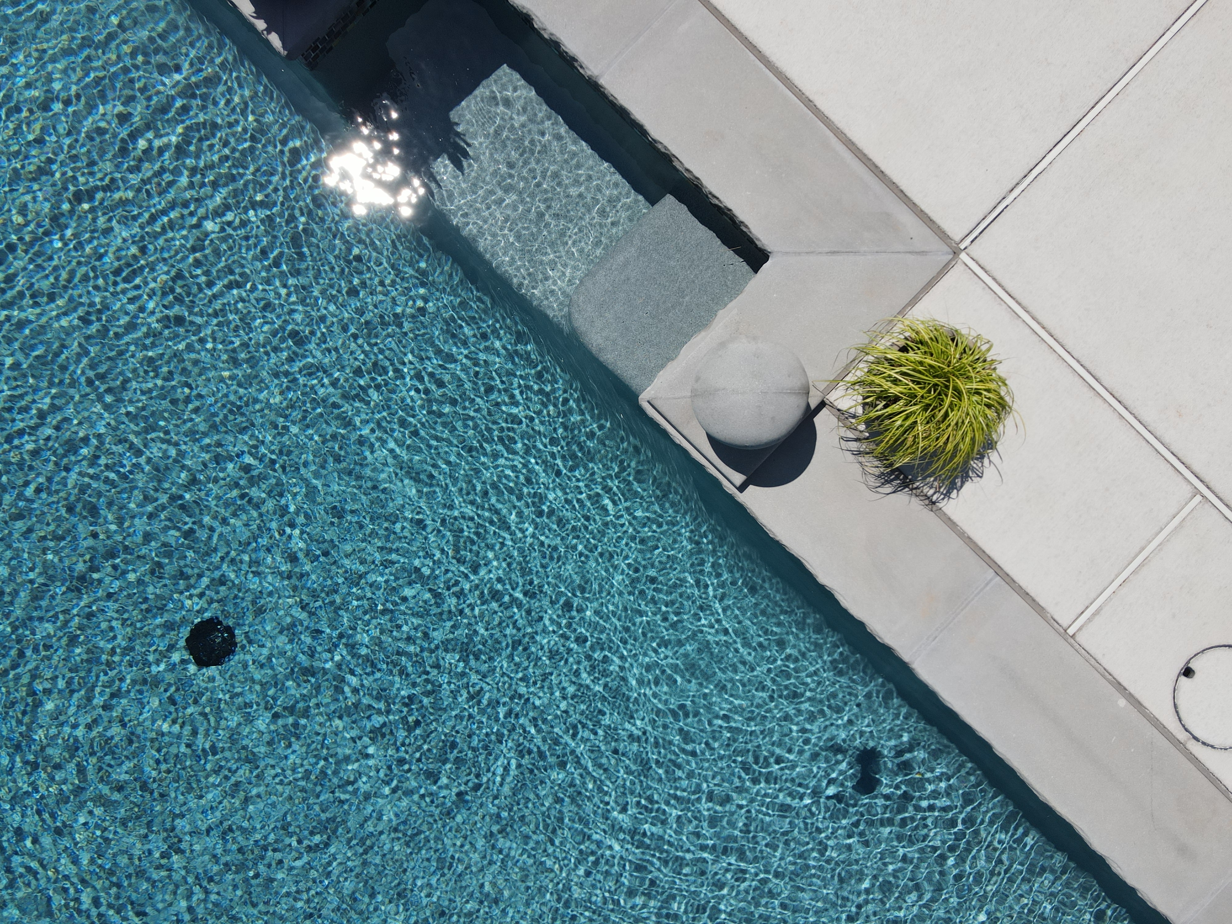 An aerial close-up of a swimming pool's edge, showing a set of submerged steps with a dark, textured finish. The deck surrounding the pool is made of light grey, square pavers. A grey sphere and a potted green plant are placed on the deck, near the pool's edge.