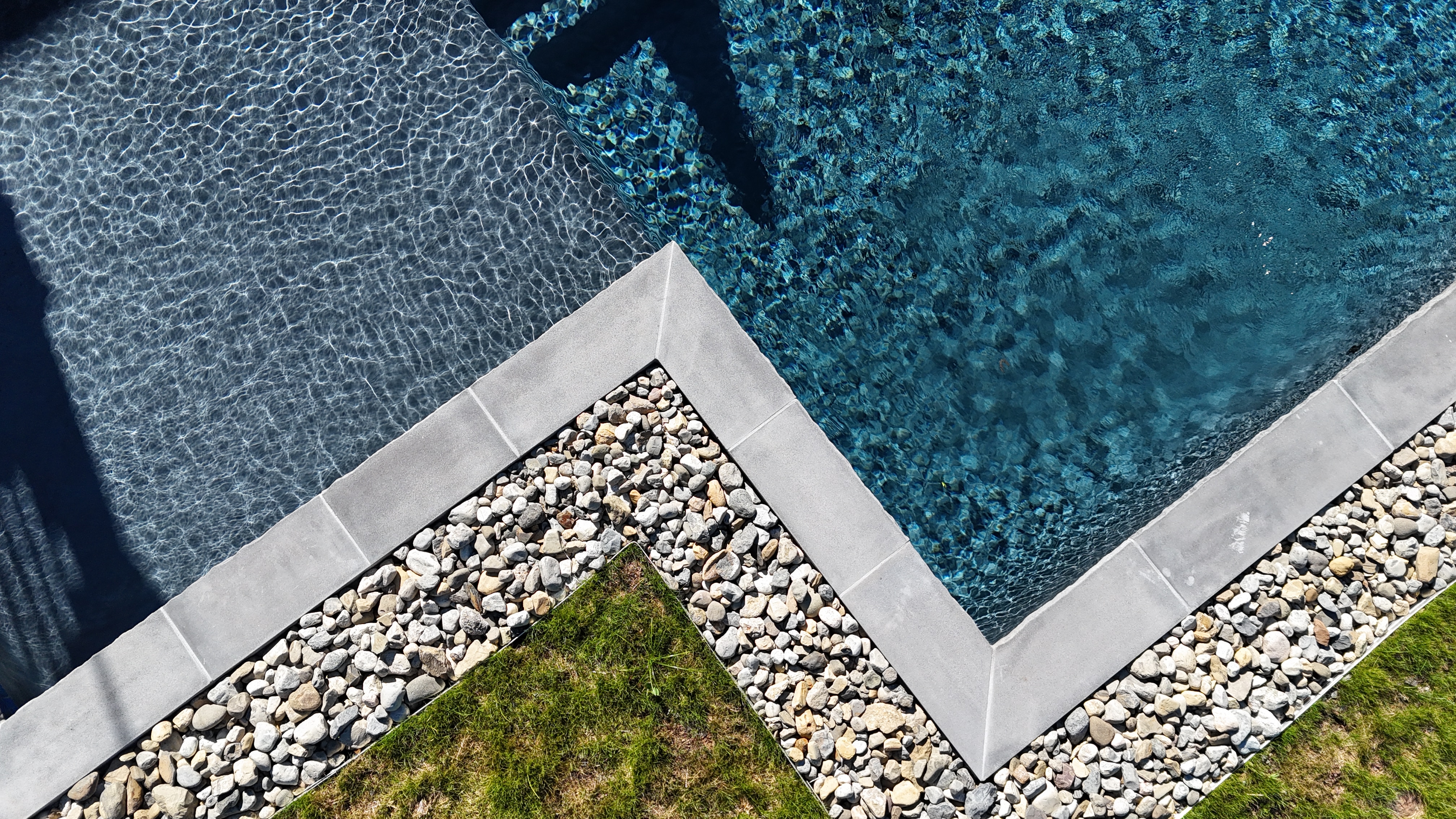 An overhead shot of the corner of a swimming pool. The pool's coping is a dark gray stone, creating a sharp angle. The pool has deep blue water. The deck area is a mix of small, light-colored river stones and a patch of green grass, creating a modern and natural look.