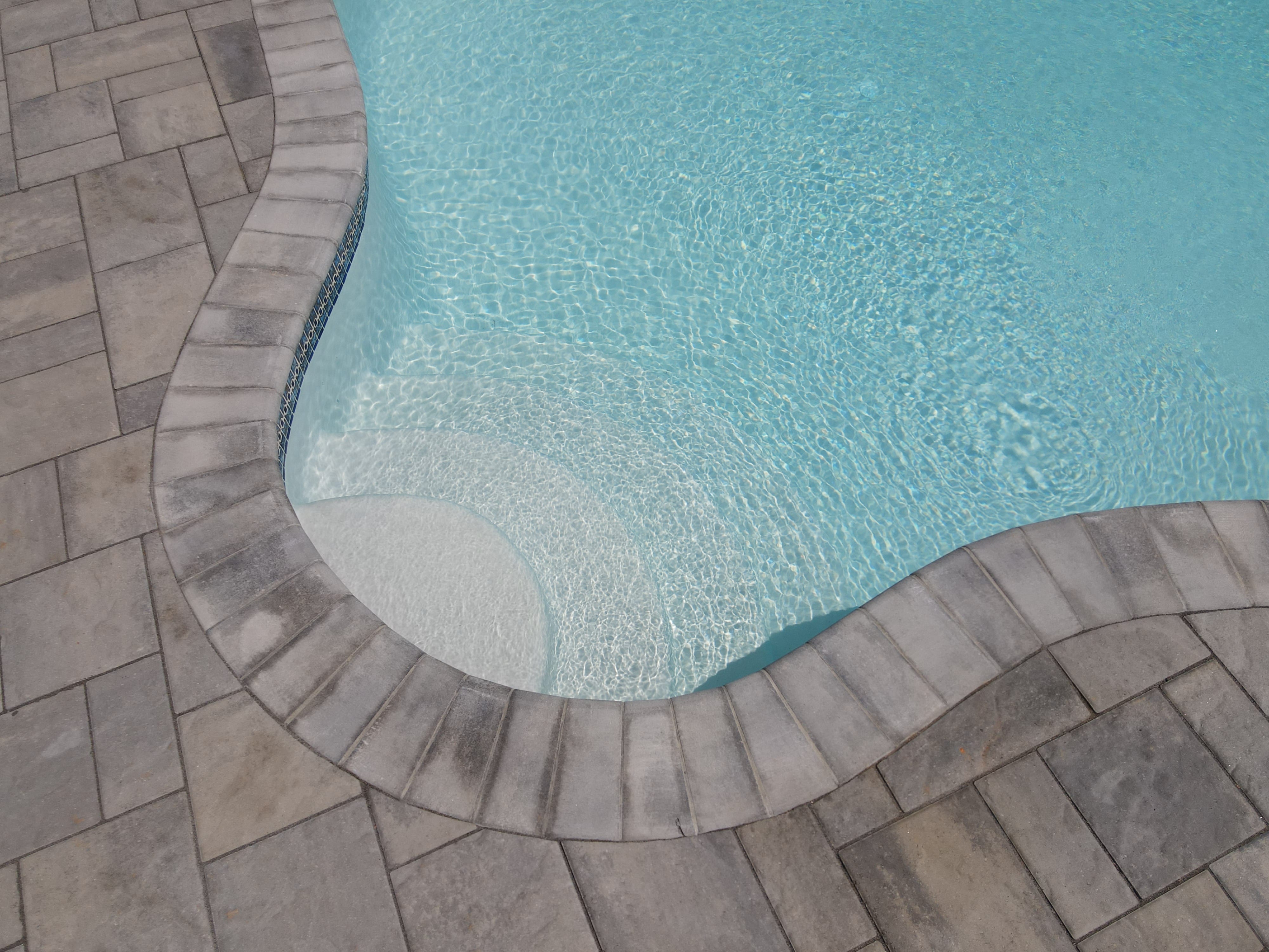 An image showing the corner of a swimming pool with steps leading into the water. The pool has a light blue interior and is surrounded by a gray paver deck. The coping, or edge, of the pool is made of larger, lighter-colored stone pavers that follow the curved shape of the pool and steps.