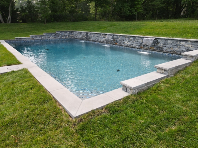 A backyard rectangular swimming pool with a stone water feature and a tiered rock wall. The pool has a light blue interior and is surrounded by a well-manicured lawn and a light gray paver deck. The coping is also a light gray stone. The stone wall features three water features from which water spills into the pool.