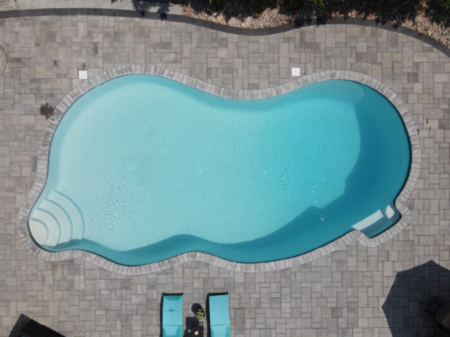 An overhead shot of a backyard with a large, free-form swimming pool. The pool has a light blue interior and is surrounded by a light grey paver patio. The patio features a circular fire pit area.