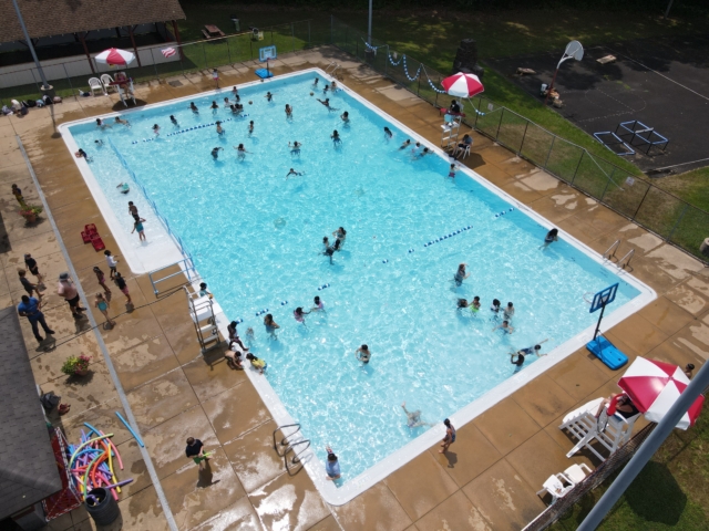 An aerial view of a rectangular community swimming pool. Many people, including adults and children, are swimming and playing in the clear blue water. A few people are sitting or standing on the concrete deck around the pool.