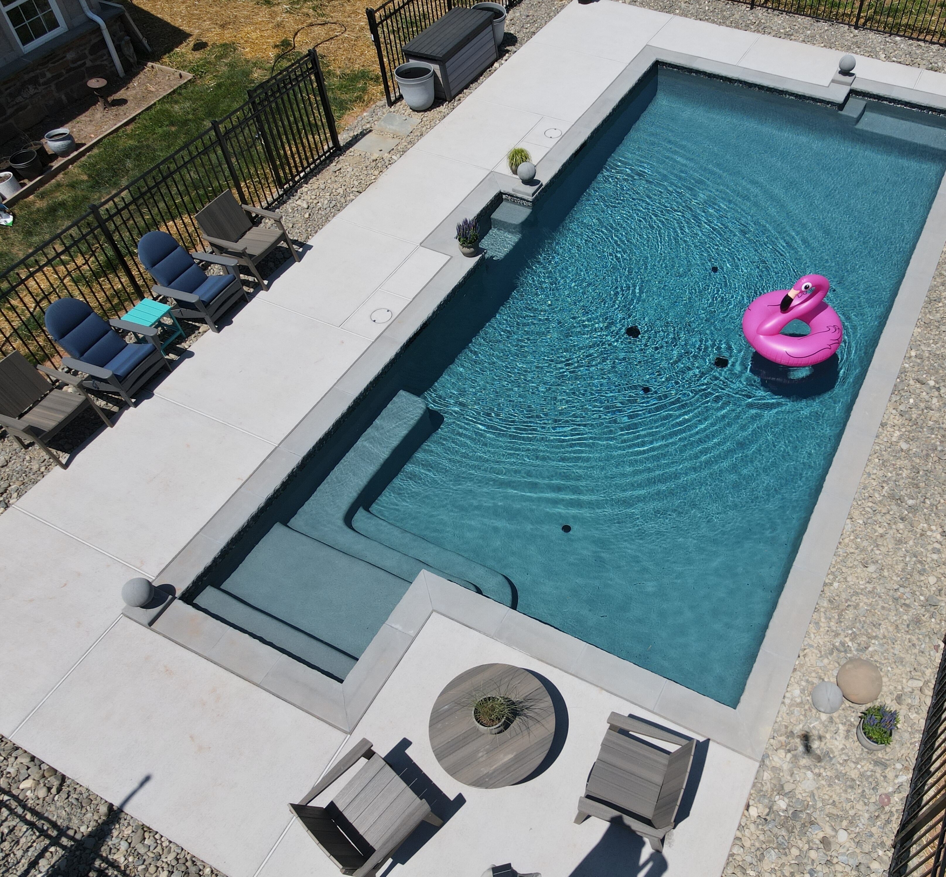 An aerial view of a rectangular, modern in-ground swimming pool with a turquoise-colored bottom. A pink flamingo float is in the center of the water. The pool has built-in stairs on one end and a small shallow area. The pool is surrounded by a light-colored concrete deck, with a small seating area and lounge chairs on one side.