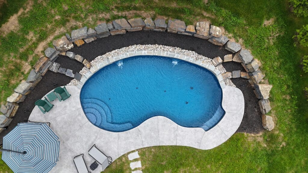 An aerial shot of a kidney shaped swimming pool with a dark blue interior. The pool is surrounded by a light-colored patio and a perimeter of gray river stones, which are bordered by dark mulch.