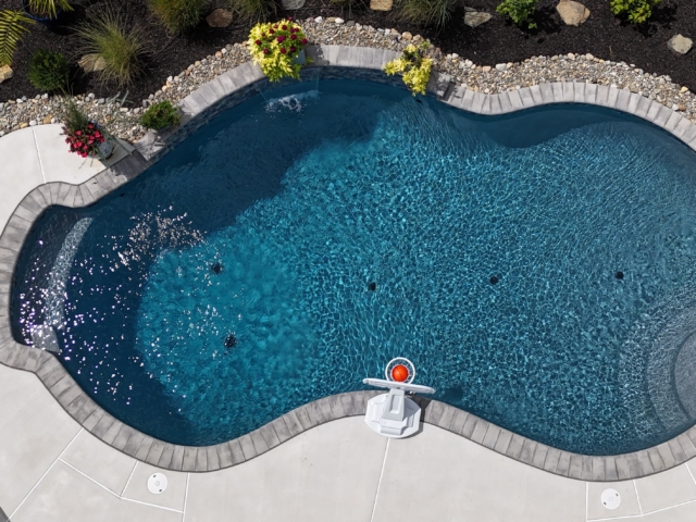 An aerial view of a custom-shaped swimming pool with a dark blue interior. The pool has a curved, irregular shape and is surrounded by a light-colored concrete deck with a decorative border of gray stones. A stainless steel hand railing with steps is also visible on the right side.