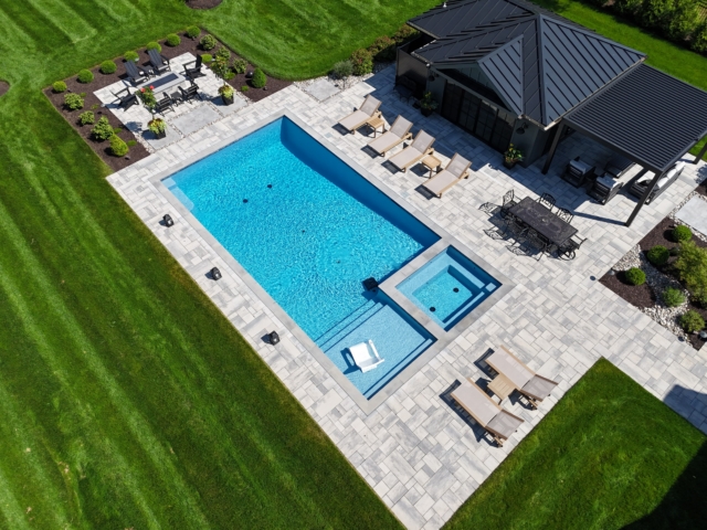 An aerial view of a luxurious backyard with a rectangular swimming pool and a smaller spa. The pool and spa are surrounded by a patio made of gray pavers. A dark-colored pool house with a black roof and a covered patio area is on the right side of the image. A set of steps leads from the pool into a shallow sun shelf with a white chair.
