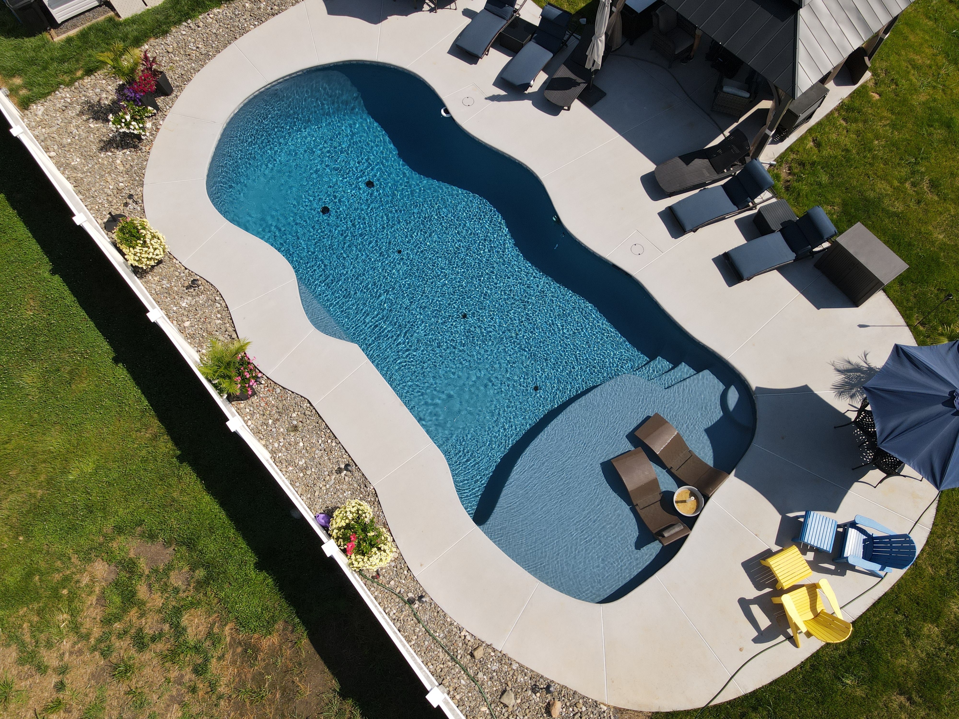 An aerial view of a backyard swimming pool with a unique, curvy shape and a sun shelf at one end. The pool has a dark blue interior and a light-colored concrete deck. On the sun shelf, there are two lounge chairs, and in the background, a few colorful Adirondack-style chairs are visible under a blue umbrella.