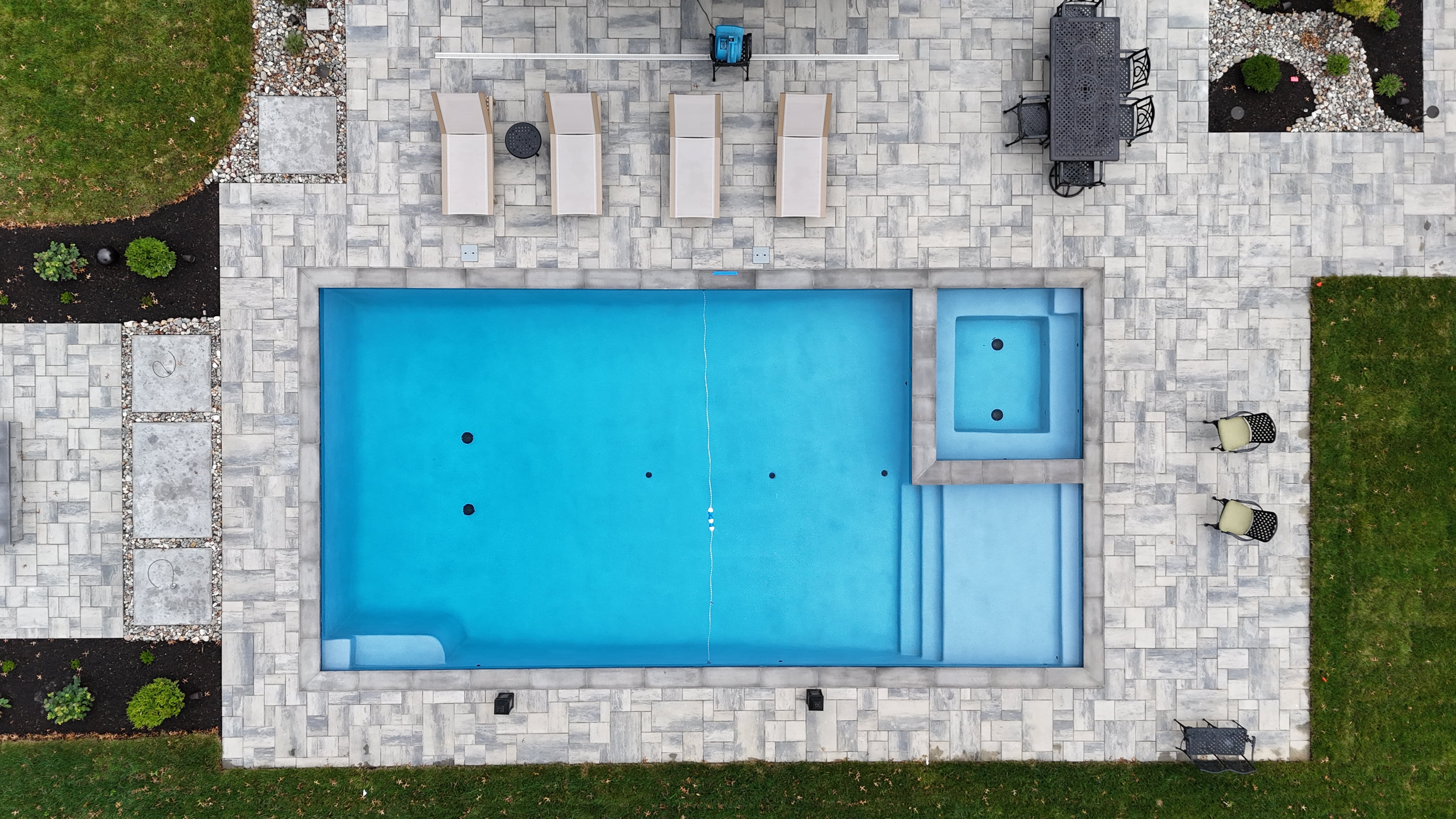 An overhead drone shot of a rectangular swimming pool and spa. The pool is filled with clear blue water and has a small sun shelf and steps at one end. A square spa is located next to the pool, separated by a small wall. The entire pool area is surrounded by a gray paver patio.