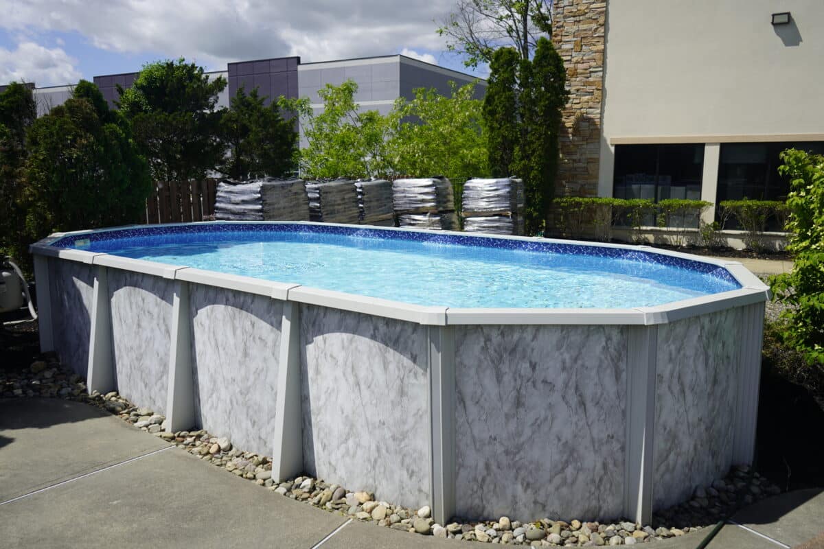 An oval-shaped, above-ground swimming pool in a display yard. The pool has a blue tiled interior and a light gray, faux-marble-textured exterior with white supports. The pool is on a concrete pad, surrounded by a building with stone siding and pallets of supplies.