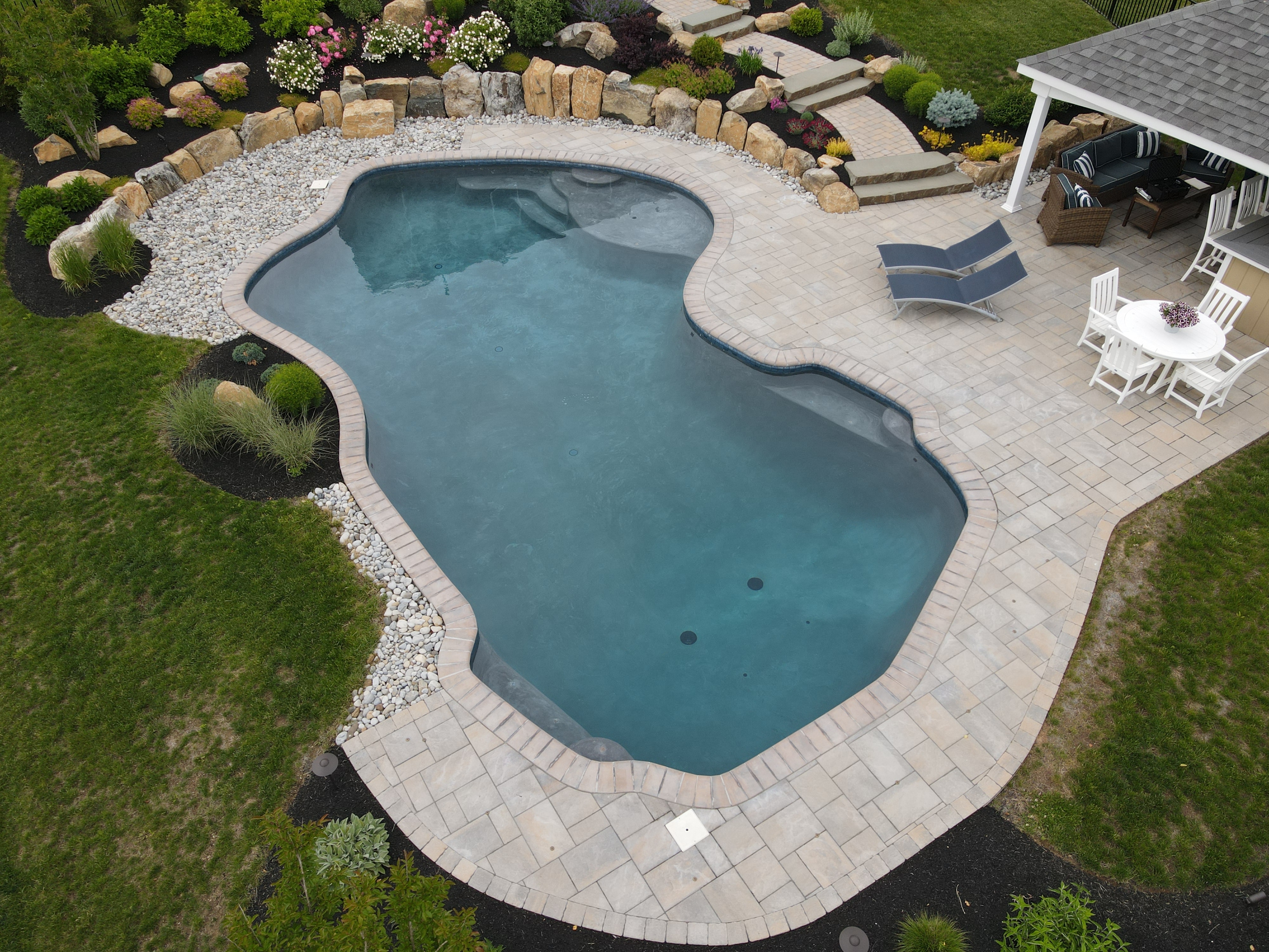 An aerial view of a backyard with a freeform swimming pool and a small, attached pergola. The pool has a dark blue interior and is surrounded by a light-colored paver deck. A set of stepping-stone stairs leads up a small, landscaped hill with a rock garden to a grassy area.