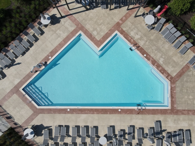 An aerial view of a geometrically shaped swimming pool with bright blue water. The pool has a large, triangular main section and a rectangular offshoot. A wide concrete deck surrounds the pool. A short staircase with a handrail leads into the pool from the shallow end. The deck has a decorative border of darker, reddish pavers.