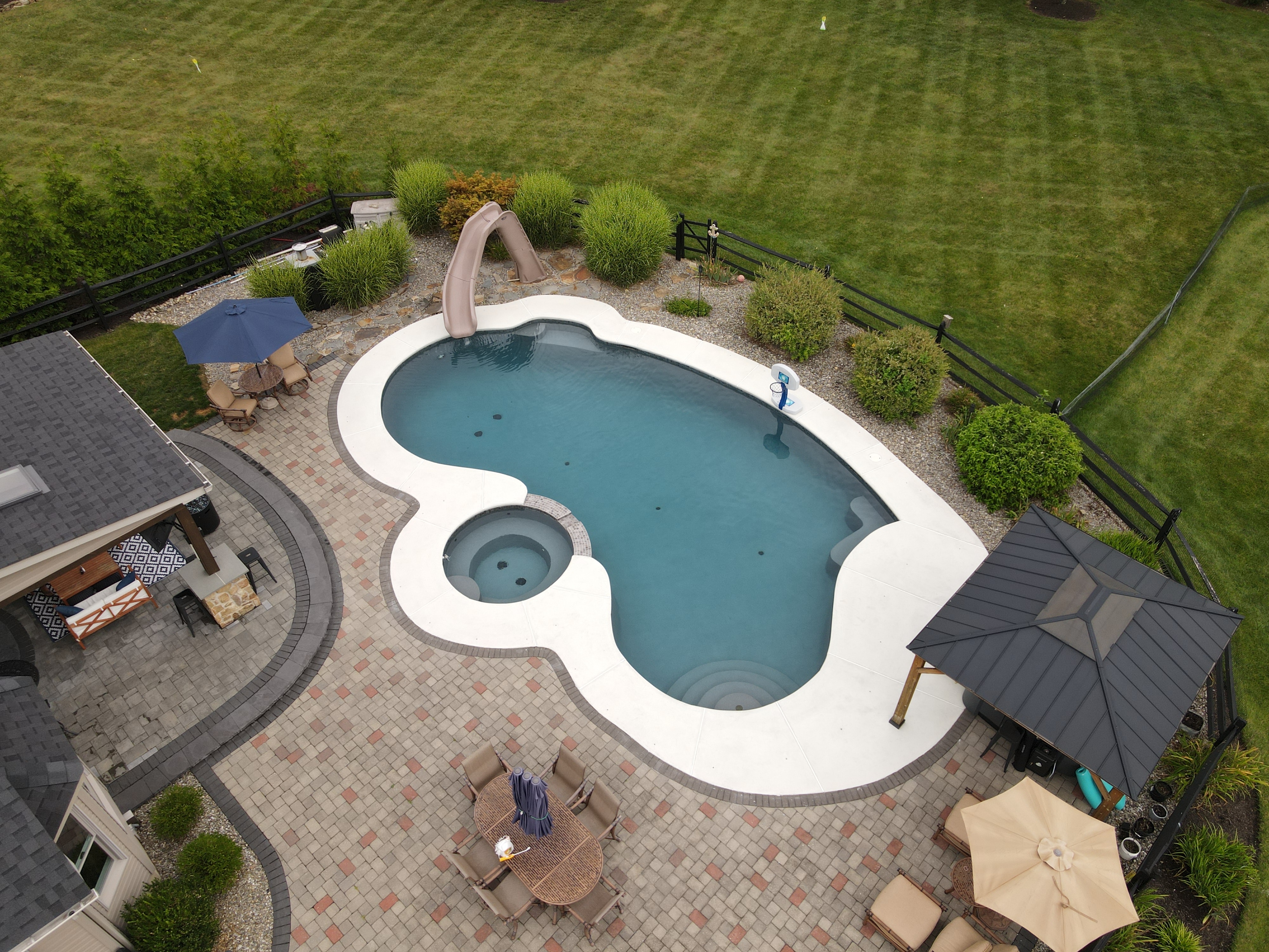 An overhead view of a large, custom-shaped backyard swimming pool with a connecting hot tub. The surrounding patio is a light concrete. The pool area features a hot tub, a built-in slide, a basketball hoop, and several seating areas, including a covered gazebo and a patio dining set.