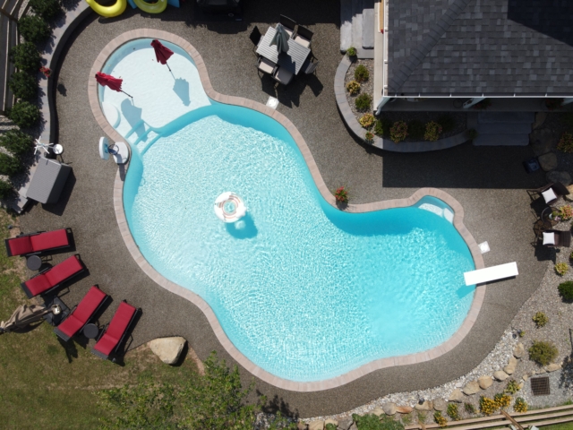 An aerial view of a custom-shaped swimming pool with a light blue interior. The pool is surrounded by a gray pebbletone deck. Two red umbrellas are placed in the shallow end of the pool. A white diving board extends from the right side of the pool, and a small, white inner tube is floating in the water.