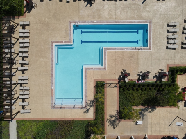 An aerial view of a large, L-shaped swimming pool with two distinct sections. The top of the 'L' is a deeper rectangular pool with two lap lanes. The longer part of the 'L' is a shallow area with steps and a handrail. The pool is surrounded by a light-colored concrete deck.