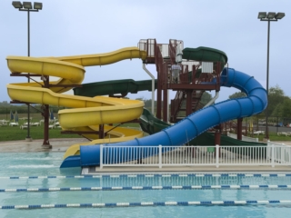 An outdoor swimming pool with a large, multi-slide water slide structure. The structure has a brown metal frame and a staircase leading to three slides: one yellow twisting slide, one green, and a blue enclosed spiral slide.