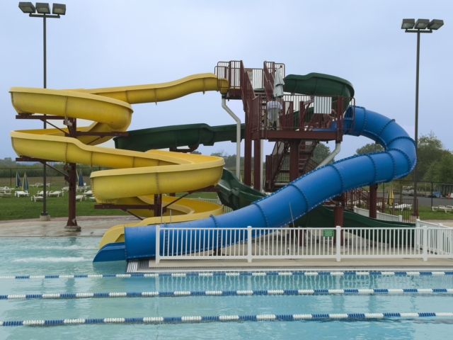 An outdoor swimming pool with a large, multi-slide water slide structure. The structure has a brown metal frame and a staircase leading to three slides: one yellow twisting slide, one green, and a blue enclosed spiral slide.